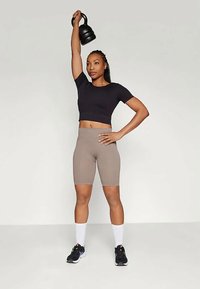 Maglietta nera corta, pantaloni da ciclismo beige a vita alta e scarpe da ginnastica nere. Il soggetto solleva un kettlebell nero sopra la testa su uno sfondo chiaro.