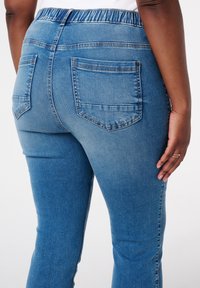 Jean en denim bleu clair avec une texture lisse, taille élastique et deux poches arrière. Présente une coupe ajustée se rétrécissant au niveau de la cheville.