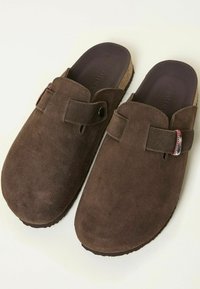 Brown suede clogs with a cork sole, featuring a decorative strap and button detail. Soft texture and slip-on design.