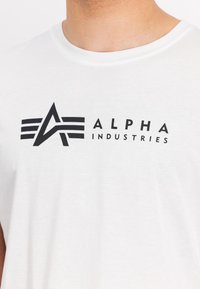 Hvid bomulds T-shirt med et sort logo med en stiliseret "A" og vandrette linjer. Rund halsudskæring, korte ærmer, minimalistisk design.