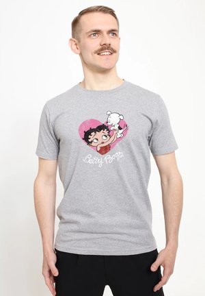 Uomo che indossa una T-shirt Betty Boop grigio chiaro con grafica di Betty Boop che tiene un piccolo cane bianco dentro un cuore rosa.