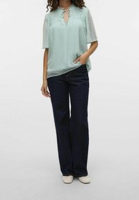 Mintgrön blus med prickar, volangkant vid halsen och genomskinliga ärmar. Kombinerad med mörkblå jeans och ljusa skor.