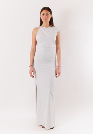 Donna in piedi di fronte a noi che indossa un lungo vestito bianco senza maniche con uno spacco laterale su uno sfondo bianco uniforme.