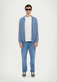 Άνδρας φορώντας μαύρα γυαλιά ηλίου, ανοιχτό μπλε φούτερ με φερμουάρ, λευκό μπλουζάκι, μπλε τζιν και γκρι αθλητικά παπούτσια, στέκεται μπροστά σε ένα απλό λευκό φόντο.