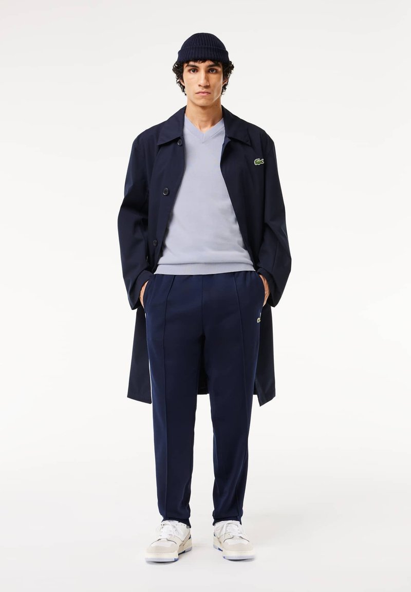 Marineblauer Trenchcoat mit Knöpfen, hellgrauer V-Ausschnitt-Pullover, passender marinefarbener Hose und weißen Sneakers. Enthält grüne Logodetails auf dem Mantel und der Hose.