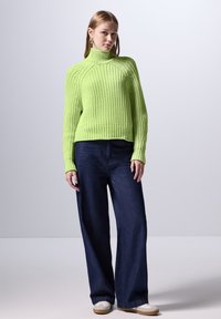 Helles grünes, strukturiertes Strick-Turtleneck-Oberteil, kombiniert mit locker sitzenden dunkelblauen Jeans und weißen Sneakers, vor einem hellen Hintergrund stehend.