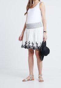 Nainen, jolla on valkoinen hihaton paita ja valkoinen hame mustalla kukkaesillä, pitää mustaa leveälieristä hattua ja käyttää hopeisia sandaaleja.