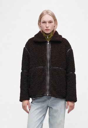 Jeune personne aux cheveux clairs portant une veste zippée marron foncé texturée sur un haut vert et un jean bleu clair, debout devant un fond blanc uni.
