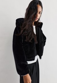Črna skrajšana jakna iz umetnega krzna s kapuco, z zadrgo spredaj in podrobnostmi na vrvici. Mehka tekstura, široki rokavi in kontrastni manšetni.