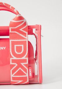 Prozoren ročni torbica z živahno koralno rožnato barvo, ki ima trak z logotipom in gladko površino; vključuje majhen notranji žep.
