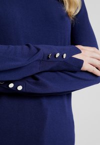 Suéter de punto azul marino con mangas largas y detalles de botones en los puños. Textura suave y diseño ajustado.