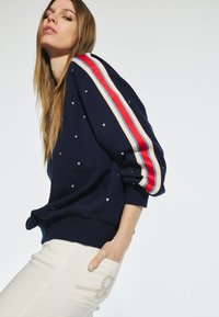 Marinblå sweatshirt med lös passform, prydd med rhinestones, har randiga röda och krämfärgade ärmar. Matchad med vita jeans och dekorativt sömnad.