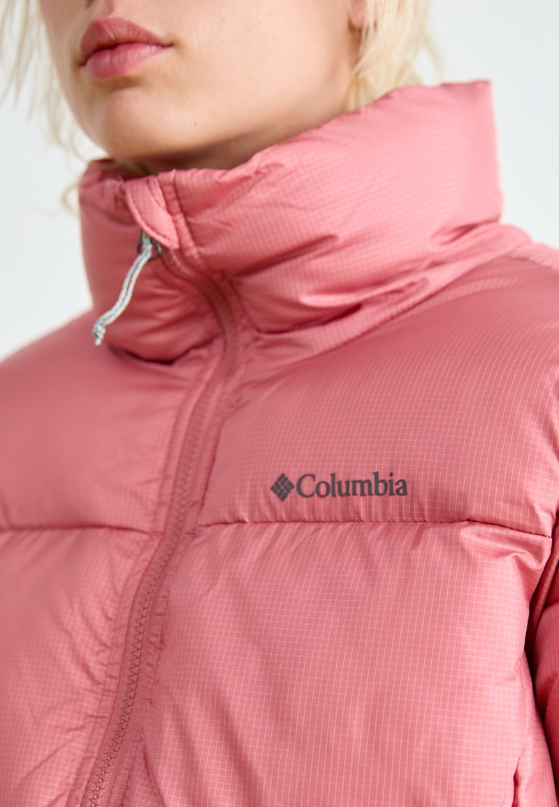 Doudoune rose clair avec col montant, dotée d'une fermeture éclair et du logo Columbia. Le tissu présente un motif à carreaux subtil et une texture douce.