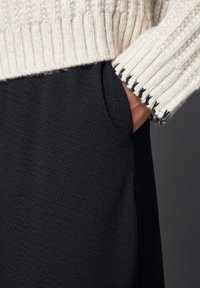 Pull en grosse maille crème avec un motif en cables et des poignets côtelés, associé à un pantalon en tissu noir texturé avec des poches latérales.