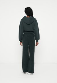 Temno zeleni hoodie z kengurujskim žepom in nastavljivo kapuco, kombiniran z širokimi, teksturiranimi temno zelenimi hlačami. Vidni so beli športni čevlji.