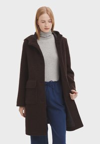 Manteau en polaire marron avec capuche, poches avant et boutons. Porté par-dessus un col roulé gris, associé à un pantalon bleu. Texture douce.