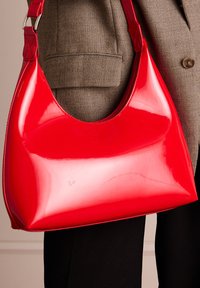 Red patent leather hobo bag with a rounded shape, flat bottom, and a single adjustable strap. Smooth, shiny surface with subtle curves.