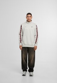 Pullover grigio chiaro con mezza zip, caratterizzato da strisce laterali rosse e nere, tasca frontale e logo Nike. Indossato con jeans scuri e sneakers.