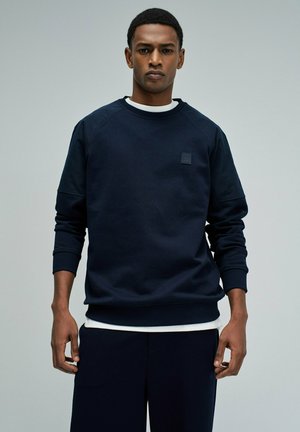 Hombre con sudadera azul marino oscuro sobre una camisa blanca, de pie frente a un fondo liso gris claro, mirando hacia adelante con una expresión neutral.