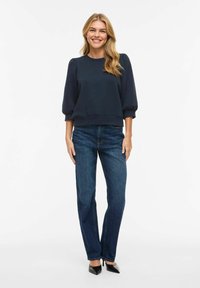 Femme debout et souriante portant un haut bleu marine à manches bouffantes, un jean bleu coupe droite et des escarpins noirs à bout pointu sur un fond blanc.