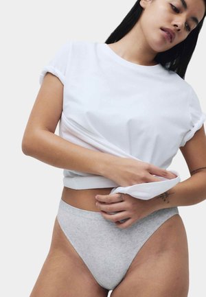 Žena zvedající bílý tričko s vyhrnutými rukávy, má na sobě světle šedé spodní prádlo s vysokým střihem a tetování na prstech a předloktí.