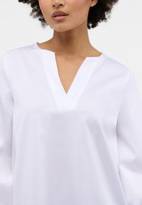 Weiße Baumwollbluse mit V-Ausschnitt und langen Ärmeln. Glatte Textur und schlichtes Design, geeignet für legere oder semi-formelle Anlässe.