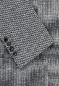 Szary materiał w wzór houndstooth o gładkiej fakturze. Posiada czarne guziki na mankietach, z widoczną kieszenią na dolnej krawędzi.
