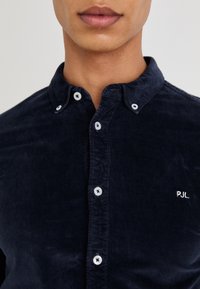 Námornícka kordová košeľa s golierom na gombíky, bielymi gombíkmi a malým vyšitým logom "PJL" na ľavej strane. Mäkká textúra.