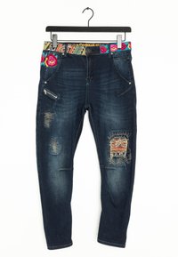 Vaqueros de mezclilla azul oscuro con una cinturilla bordada colorida, que presentan parches desgastados, cremalleras y detalles de costura ornamentales.