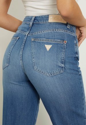 Modré džíny z denimu s vysokým pasem, klasickým pěti kapsovým designem, trojúhelníkovou nášivkou na zadní straně a jemným vyblednutím, které mají hladkou texturu.