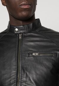 Sort læderjakke med opretstående krave, lynlåsføring foran og lille brystlomme; har en glat tekstur og minimale hardware-accenter.