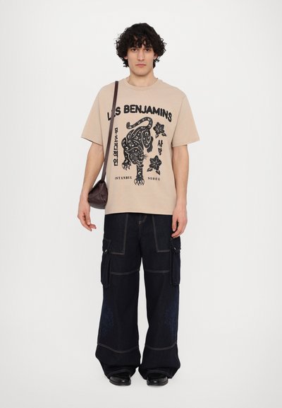 T-shirt graphique beige avec impression noire d'un panthère et de motifs floraux. Associé à un pantalon cargo bleu foncé avec des poches latérales et des jambes larges.