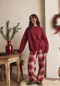 Merpunaisen sävyn collegepaita roikkuvilla olkapäillä, yhdistetty punaisen ja beigen ruutukuosisiin housuihin. Malle seisoo maljakon ja rustiikkisen pöydän vieressä.