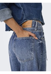 Blå denimjeans med hög midja, ljus tvätt, en läderetikett som säger "blue denim" och en bakficka med en hand som vilar inuti.