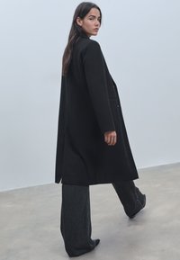 Pitkä musta takki räätälöidyllä muotoilulla, jossa on yksi nappi ja sivutaskut. Sen päällä on harmaat housut, joissa on sileä pinta.