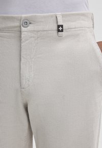 Pantaloni in corduroy grigio chiaro con una texture a coste, chiusura con bottone e un accento del marchio nero a contrasto vicino alla vita.
