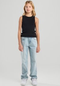 Young person standing, wearing sleeveless black top, light blue jeans, and white sneakers against a plain light grey background.