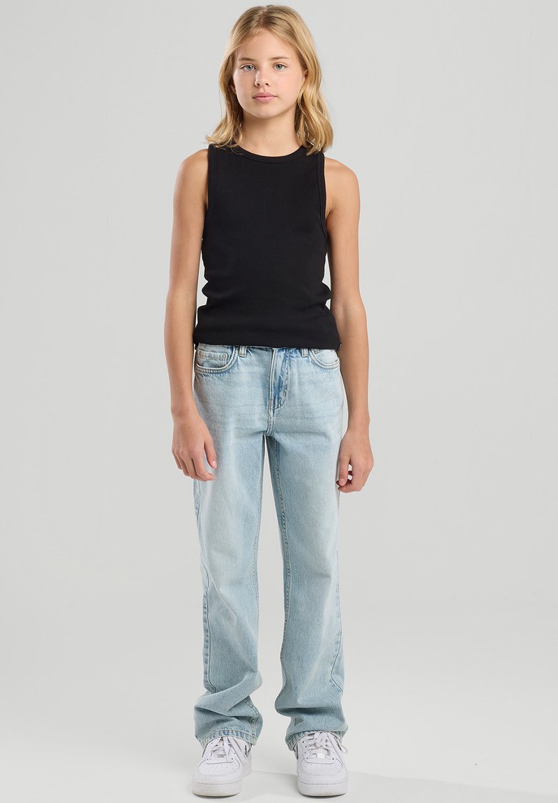 Young person standing, wearing sleeveless black top, light blue jeans, and white sneakers against a plain light grey background.