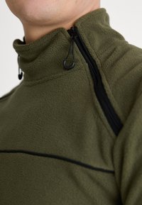 Olivově zelený fleece svetrový krk s vysokým límcem, s černým zipem na levé straně a texturovanými látkovými akcenty.