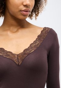 Γυναίκα που φοράει καφέ μακρυμάνικη μπλούζα με V-λαιμόκοψη διακοσμημένη με δαντέλα με λουλούδια, εμφανιζόμενη από τους ώμους μέχρι τα χείλη.