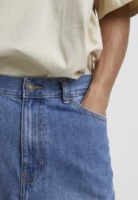 Svijetlosmeđi T-shirt je ubrizgan u plave traperice s kopčanjem na gumbe, vidljiva je prednja džepna i suptilni detalji šivanja.