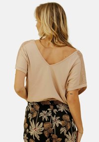 Haut à manches courtes en V de couleur beige clair avec des accents texturés sur les épaules, associé à un pantalon noir avec un motif de feuilles de palmier blanches.