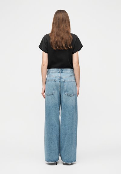 Άτομο με μακριά καστανά μαλλιά φοράει μαύρο τοπ με κοντά μανίκια και ανοιχτό μπλε τζιν παντελόνι ψηλόμεσο, με φαρδύ πόδι, στέκεται γυρισμένο προς τα πίσω σε απλό φόντο.