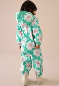 Mintgrüner wasserdichter Overall mit großen weißen Gänseblümchen und pinken Mittelpunkten, elastischen Bündchen und Kapuze, kombiniert mit weißen Stiefeln.