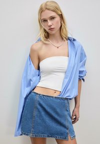 Ung kvinde iført en hvid strapless top, en uknappet blå stribet skjorte og en denim mini nederdel, stående mod en simpel lys væg.