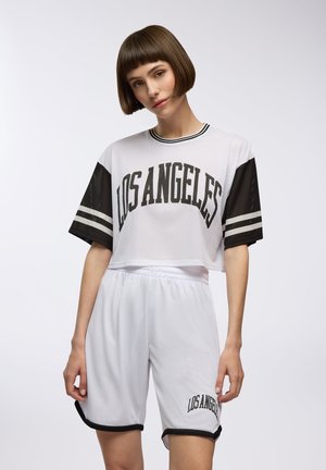 Top corto bianco in rete con maniche nere, con la scritta "LOS ANGELES" in grande lettering nero. Abbinato a pantaloni corti bianchi e dettagli neri.