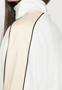 Veste blanche à col montant avec un panneau beige texturé et un passepoil noir le long de la couture, affichant un design minimaliste.