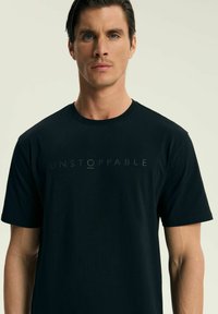 Czarny bawełniany t-shirt z krótkim rękawem, z subtelnie wytłoczonym napisem "UNSTOPPABLE" na piersi. Gładka faktura, standardowy krój.