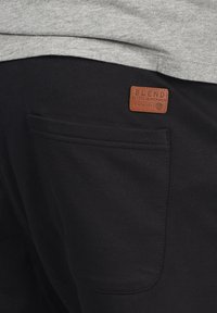 Pantalones de chándal negros hechos de una tela suave, con un parche rectangular de cuero marrón etiquetado "BLEND" y un bolsillo trasero con detalles de costura.