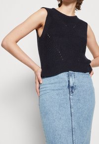 Mörkblå, ärmlös stickad tröja med ribbad textur och diagonala sömmar. Bärs över en ljusblå denimkjol.